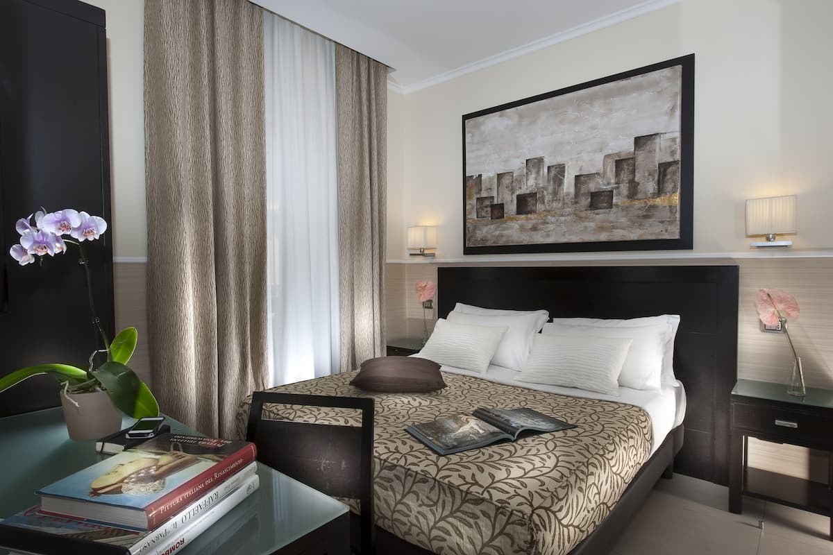 A hotel room with a bed, a desk with a lamp, a vase with flowers, and a painting on the wall.