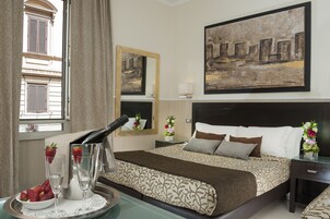 A hotel room with a bed, a bedside table, a nightstand, a window with curtains, a wall-mounted painting, and a tray with strawberries and champagne glasses.