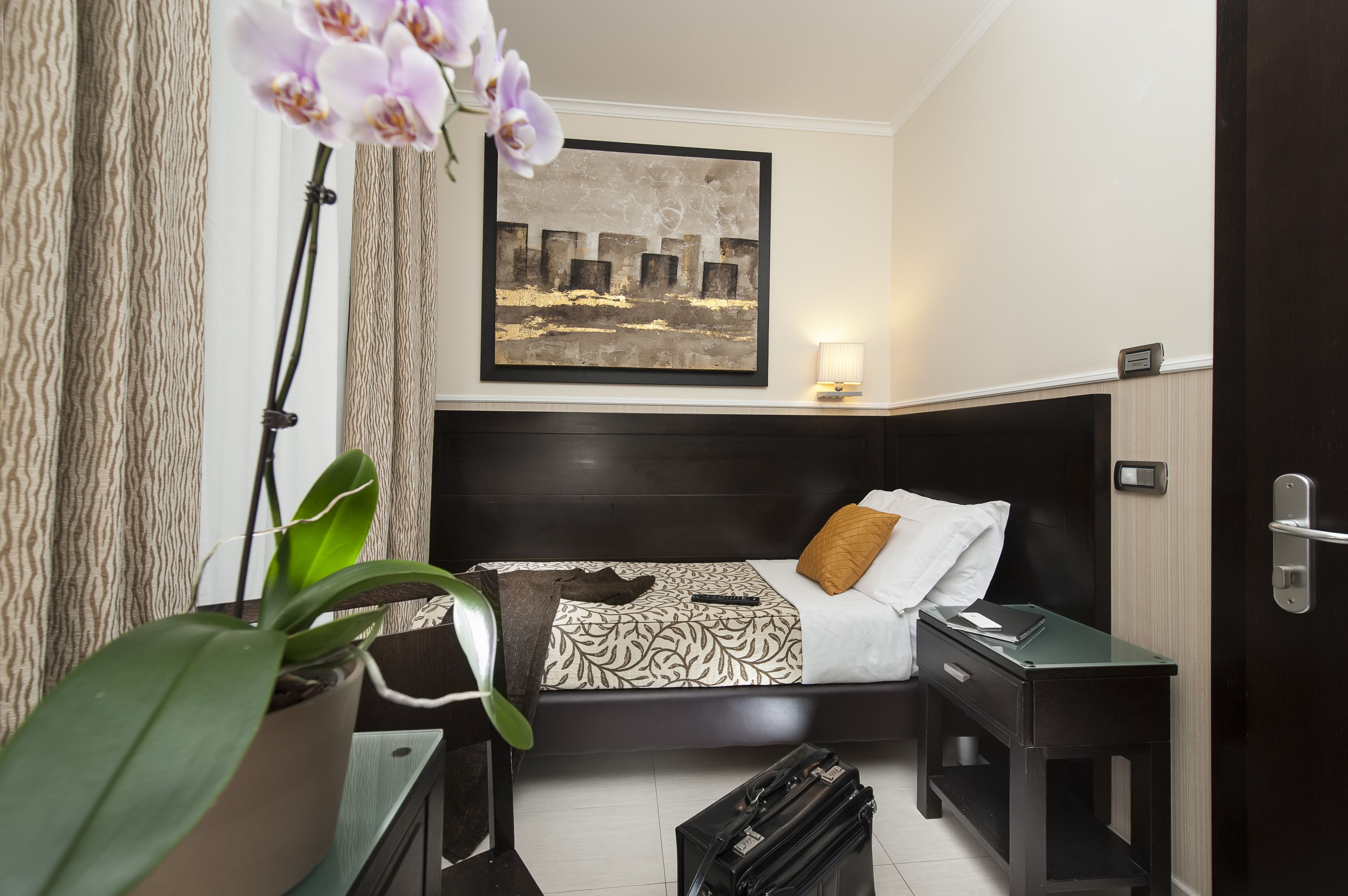 A hotel room with a bed, a nightstand, a desk, a potted plant, and a painting on the wall.