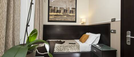 A hotel room with a bed, a nightstand, a desk, a potted plant, and a painting on the wall.