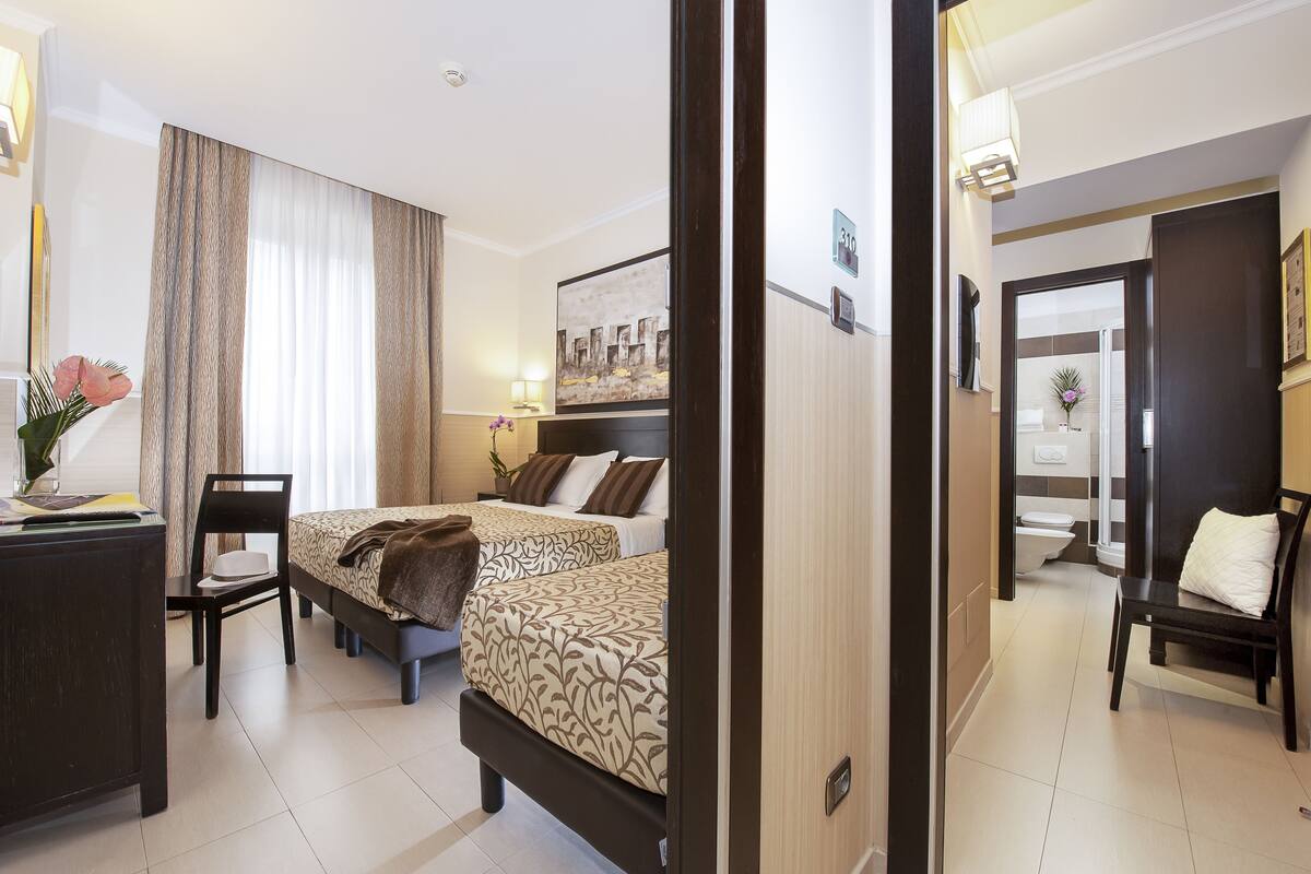 A hotel room with two beds, a desk, a chair, a bedside table, and a bathroom visible through an open door.