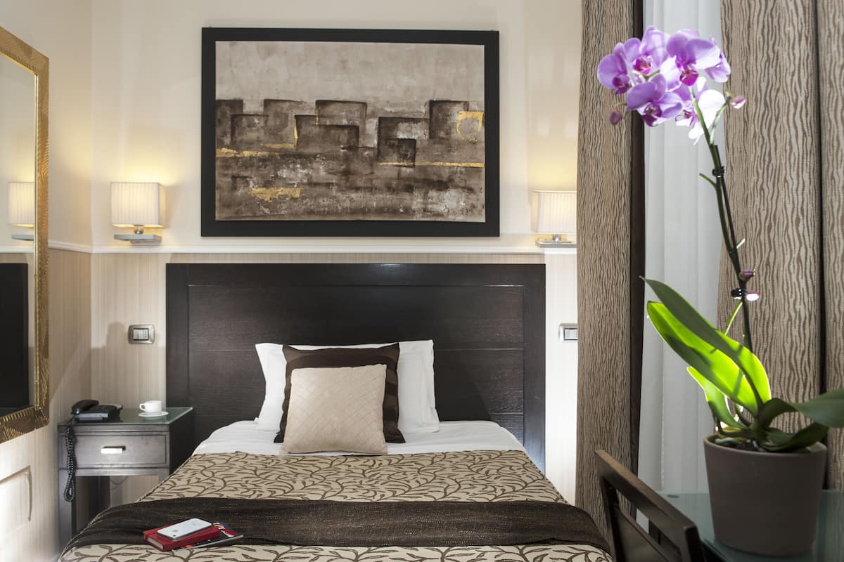 A hotel room with a bed, a bedside table with a lamp, a telephone, a cup, a book, a large abstract painting, and a potted orchid.