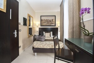 A hotel room with a bed, a desk, a chair, a potted plant, and a painting on the wall.