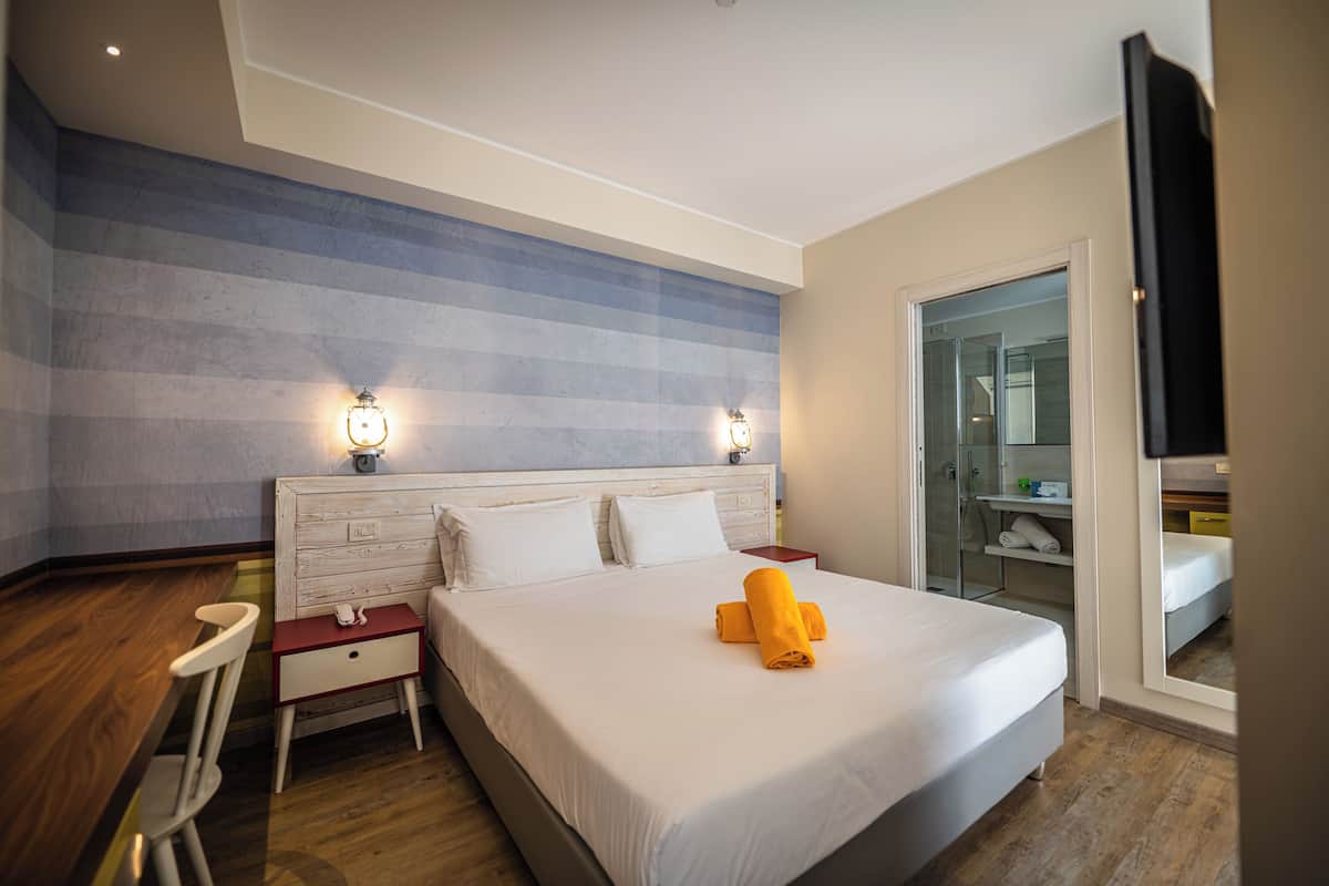 Hotelkamer met een groot bed, een houten bureau, een stoel, een nachtkastje en een douchecabine met glazen wanden.