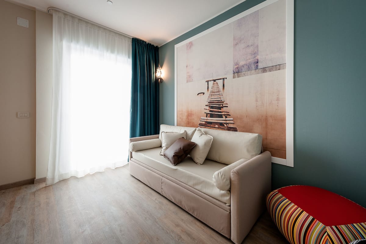 Een woonkamer met een beige bank, een rode poef, een groot abstract schilderij en een raam met transparante gordijnen.