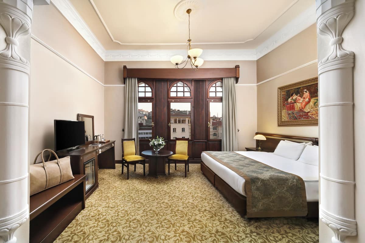 Quarto de hotel com uma cama, uma secretária com televisão, uma cadeira, uma mesa com um vaso e uma janela com cortinas.