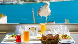 Una mesa con alimentos para el desayuno y una gaviota en un balcón con vistas a la ciudad y al mar azul.