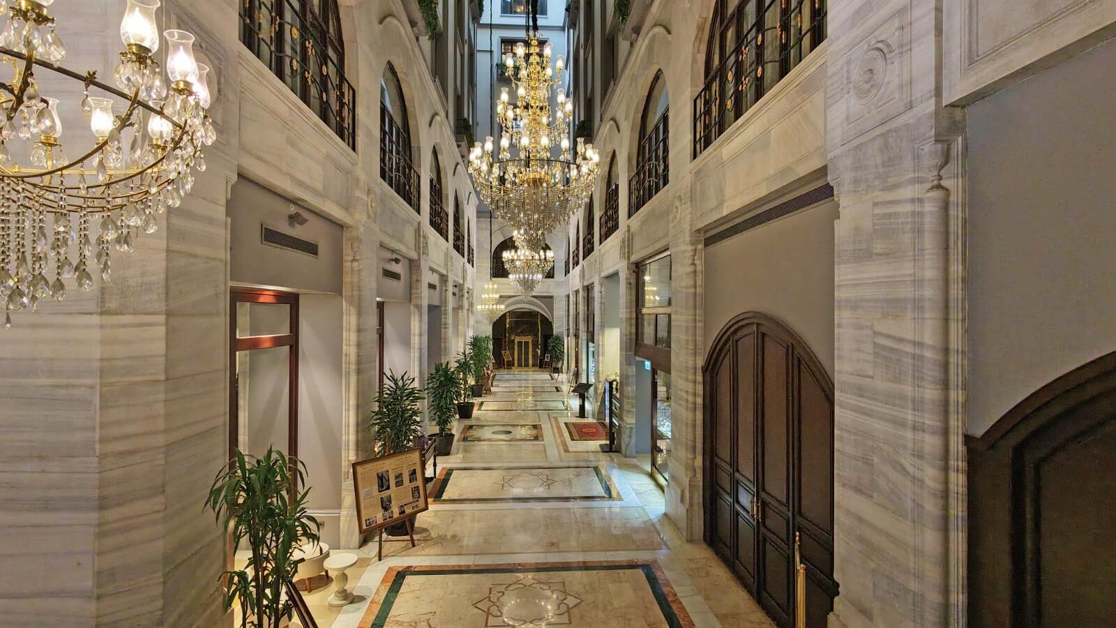 A grand hotel lobby with a high ceiling, chandeliers, marble floors, and a central staircase.