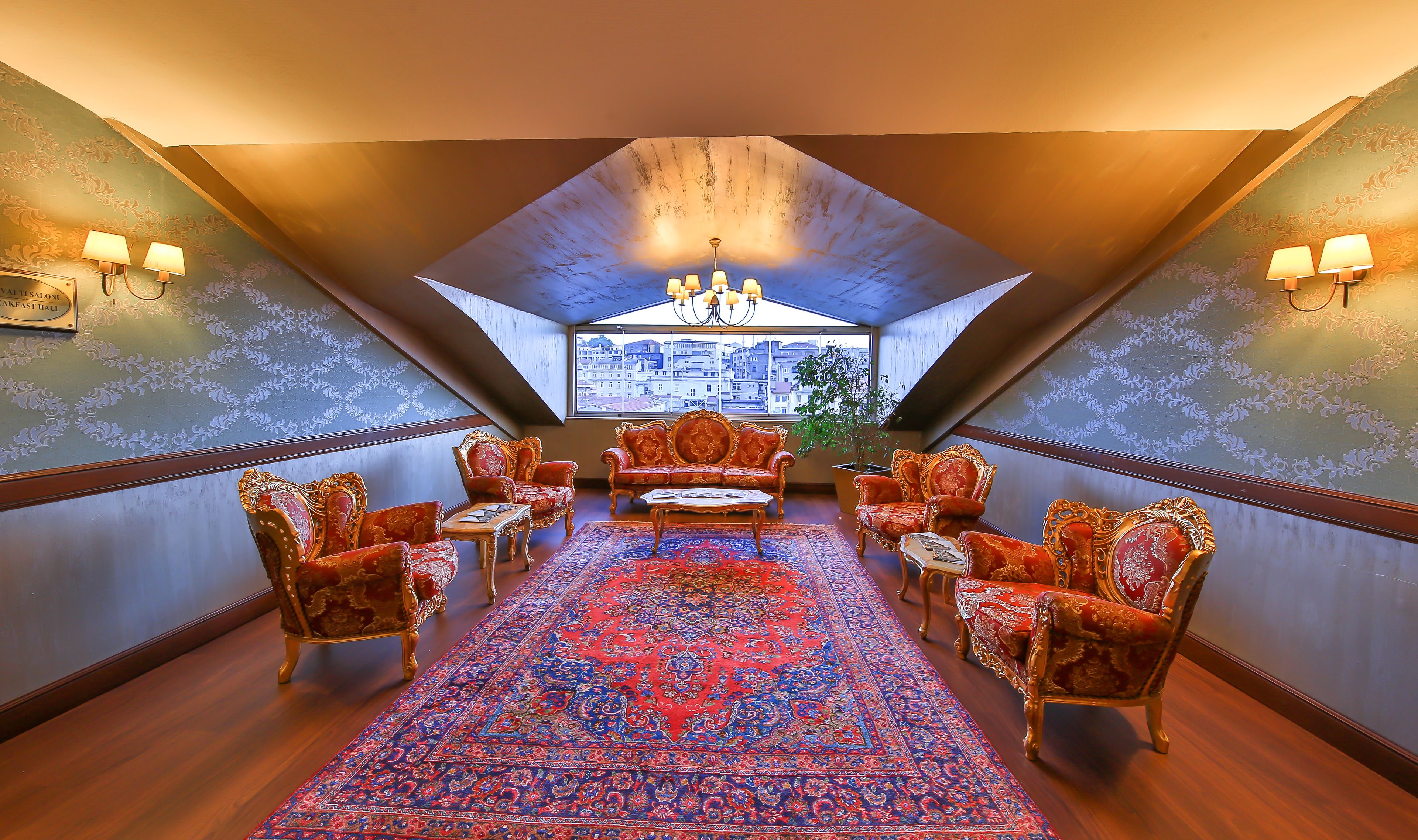 A spacious room with a high ceiling, ornate chairs, a patterned rug, and a view of buildings through a large window.