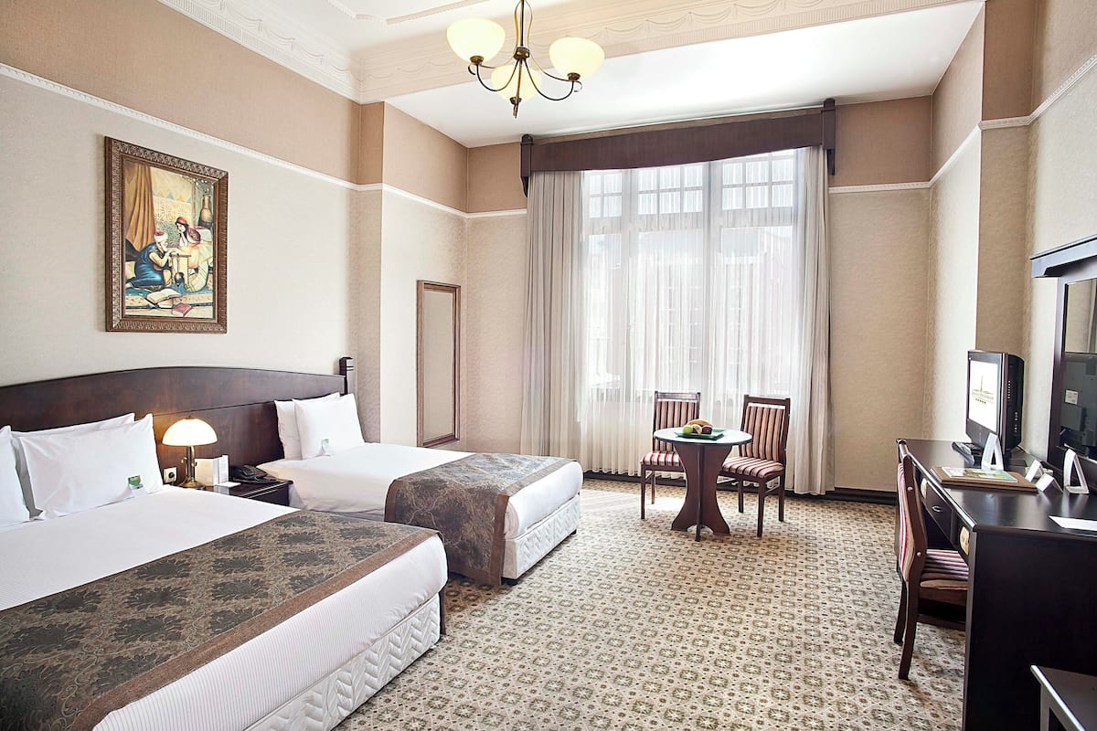 A hotel room with two beds, a desk, a chair, a television, and a large window with curtains.