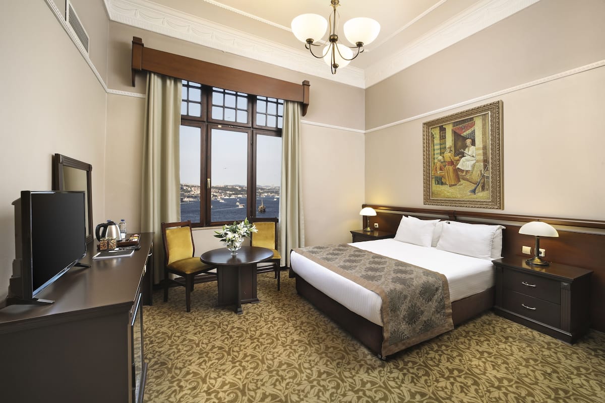 A hotel room with a bed, desk, chair, and a large window offering a city view.