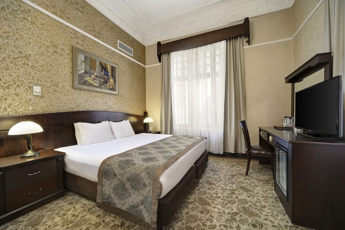 A hotel room with a large bed, a desk with a television, a chair, and a lamp.