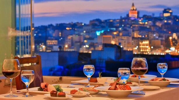 Mesa lista para la cena con varias copas de vino y platos de comida, con vistas a un paisaje urbano al anochecer.