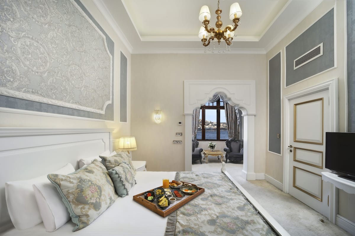 A hotel room with a bed, a tray of breakfast, a chandelier, and a view of the outside.