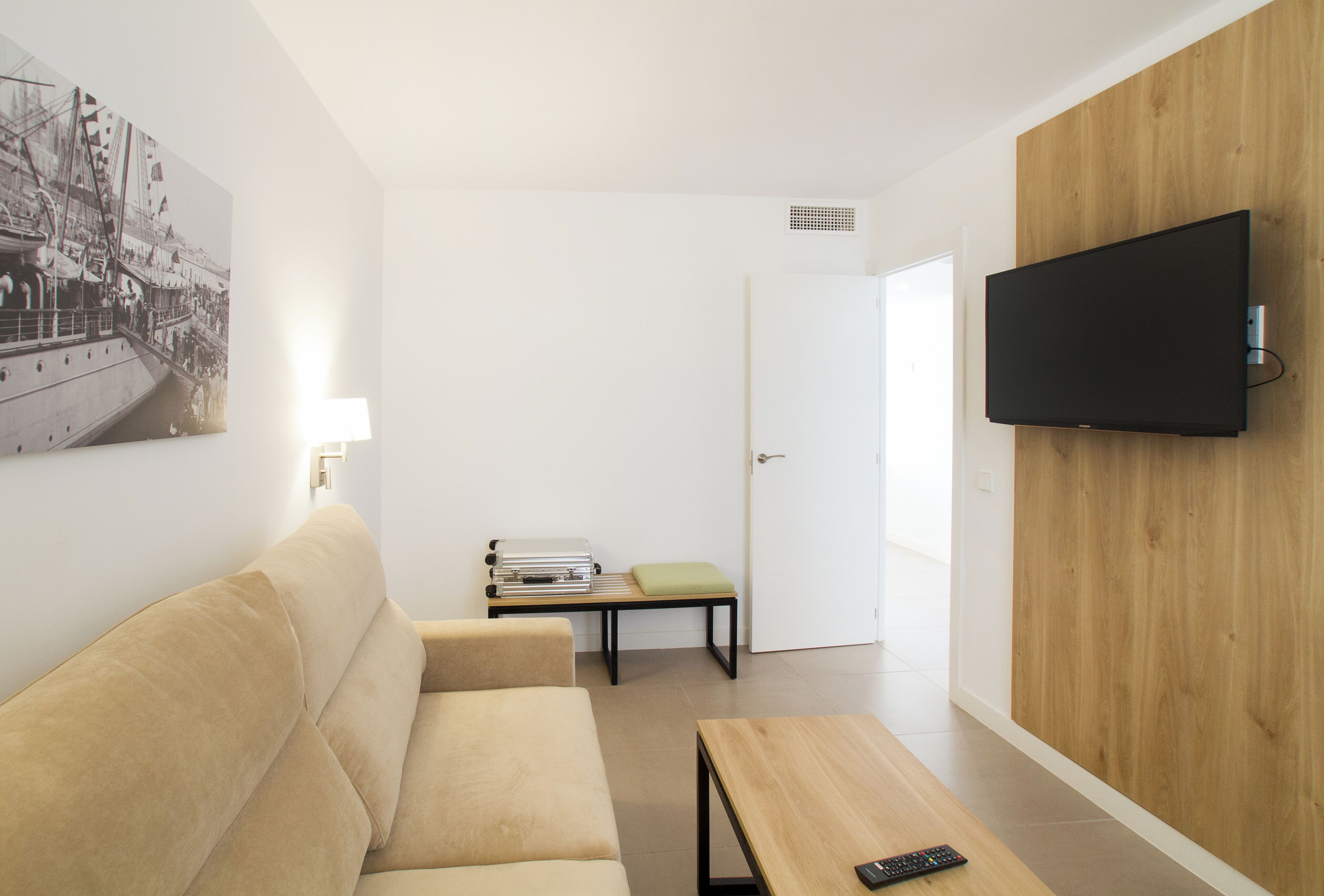 A modern living room with a sofa, a coffee table, a TV mounted on a wooden wall, a bench, and a door leading to another room.