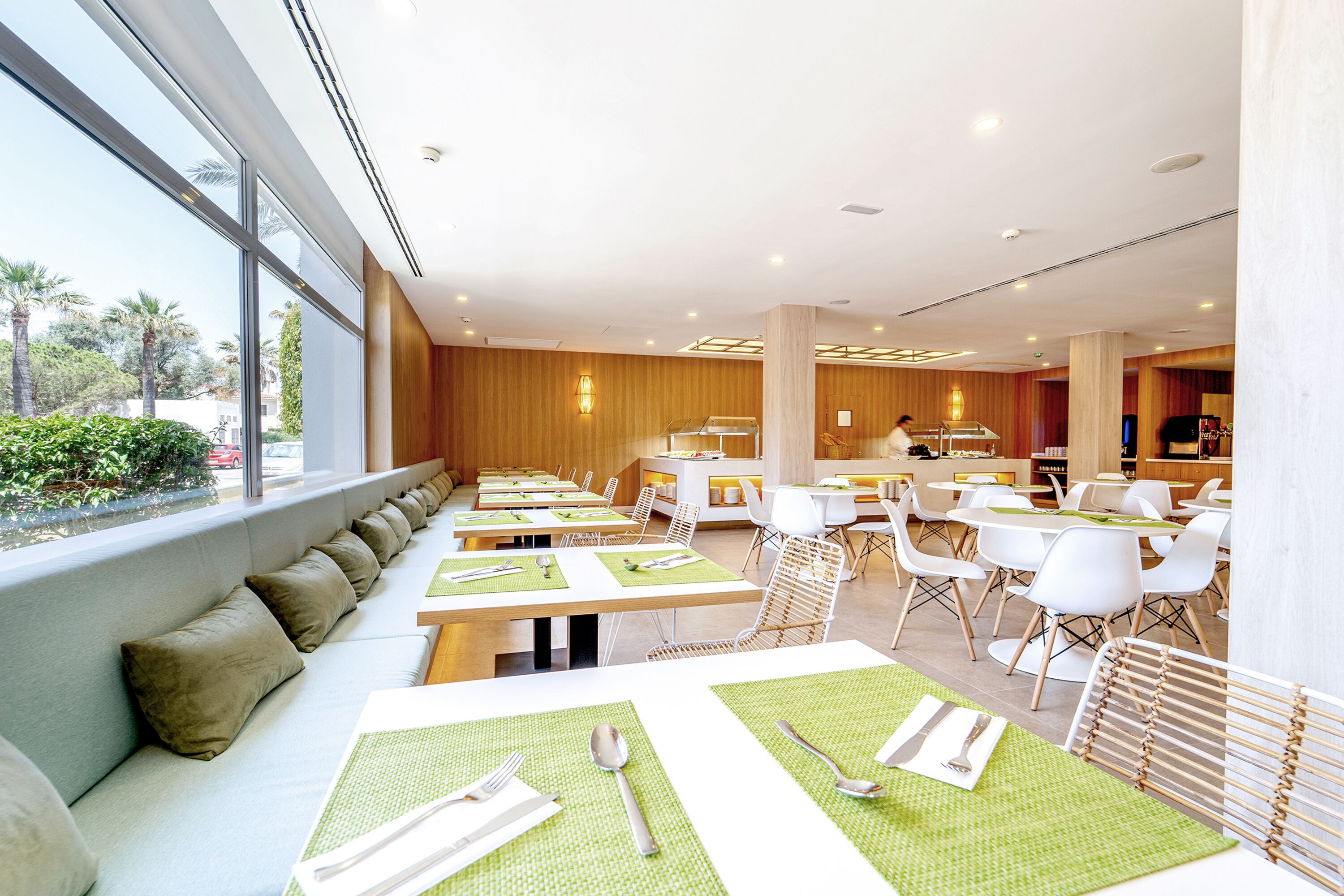 A modern dining area with long benches and tables, a kitchen in the background, and large windows offering a view of the outdoors.