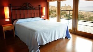 A bedroom with a large bed, wooden floor, and a city view through large windows. - Triskel (Sos del Rey Catolico)