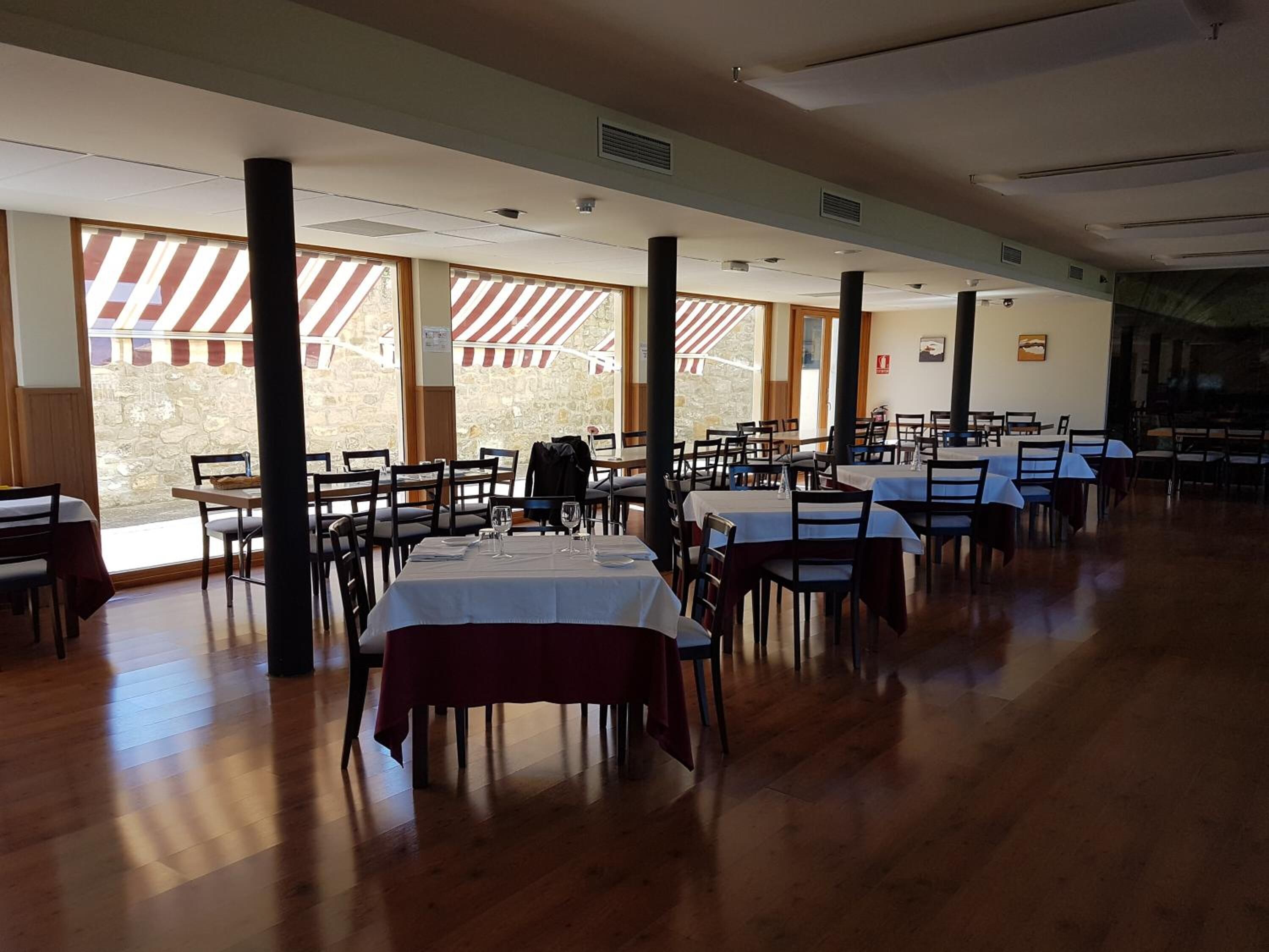 Un vaste espace dédié à la salle à manger, avec de nombreuses tables et chaises, de grandes fenêtres surmontées d’auvents à rayures et un mur en pierre en arrière-plan.