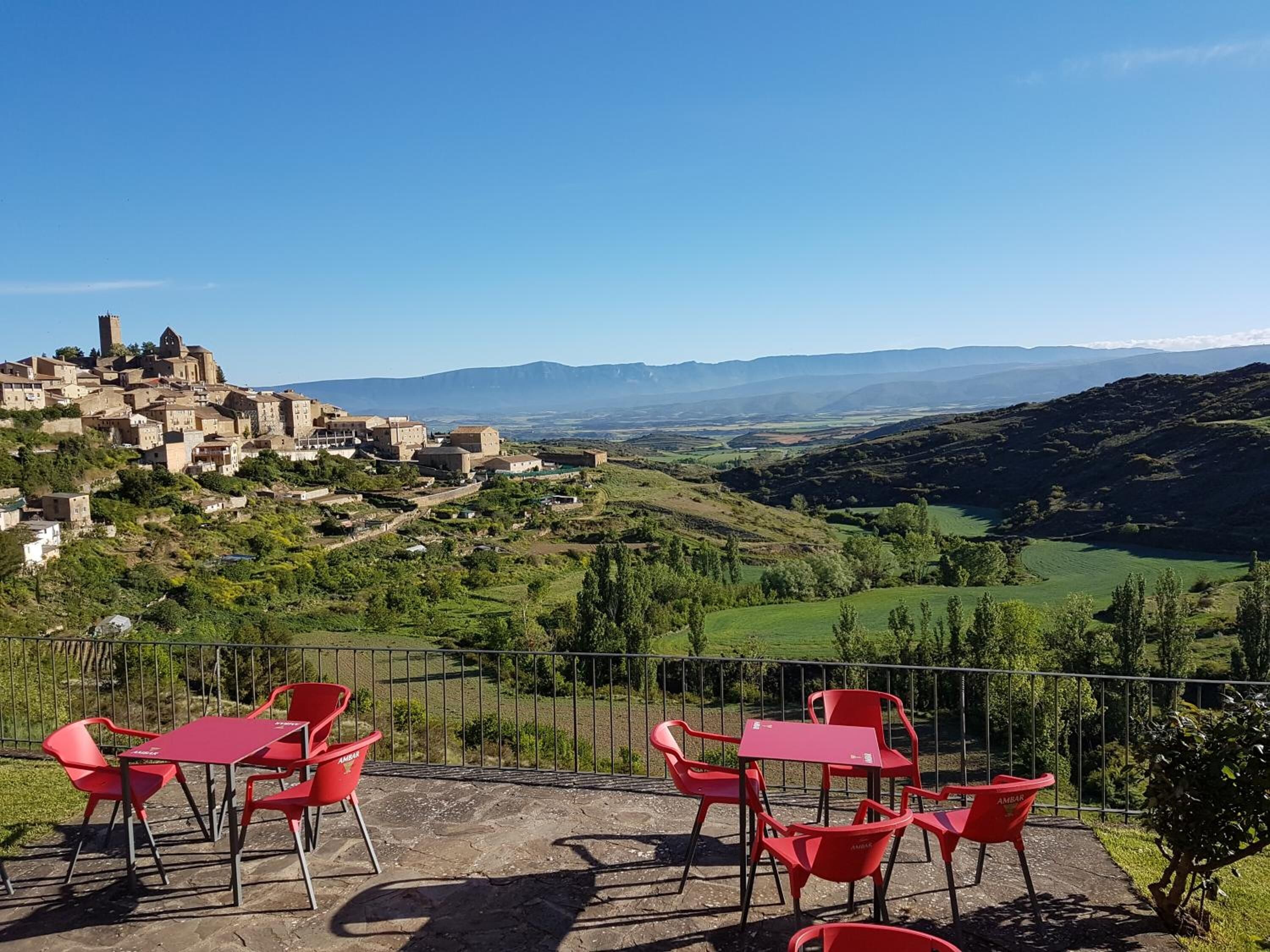 Una terraza con sillas y mesas rojas que da a un pueblo histórico con una torre destacada.