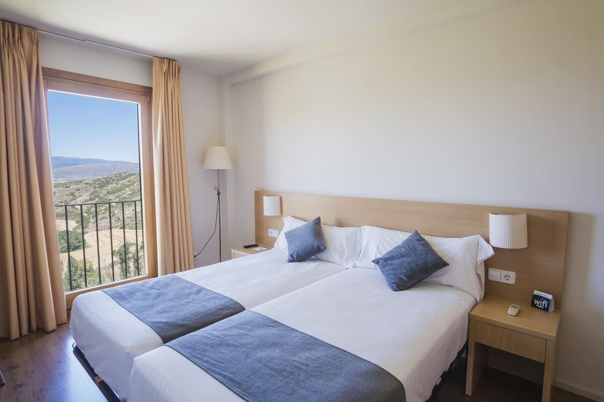 A double bed with two pillows, a bedside table with a lamp, and a view of a landscape through a window with curtains.