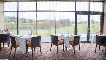 A dining area with tables set for four, large windows with a view of a green landscape, and a patterned carpet.
