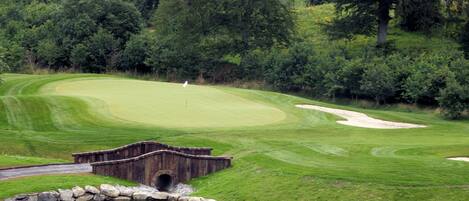 A golf course with a wooden bridge over a pond, surrounded by trees and a well-maintained lawn.