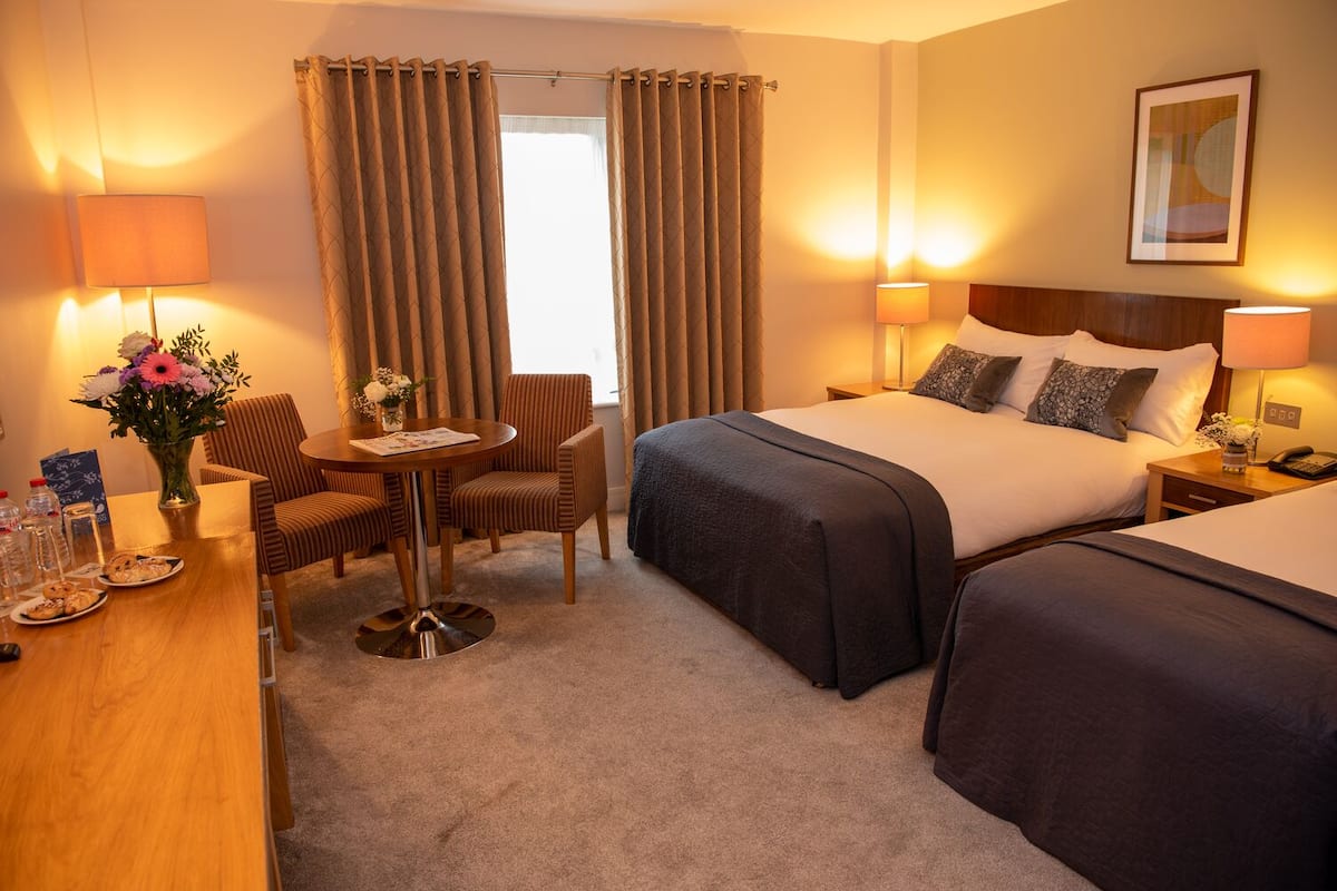 Quarto de hotel com cama, duas mesas de cabeceira, uma cadeira, uma mesinha com um vaso de flores e uma janela com cortinas.