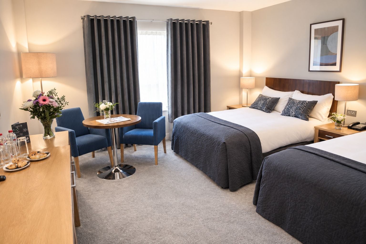 Quarto de hotel com cama, duas cadeiras, mesa com um vaso de flores e abajur.