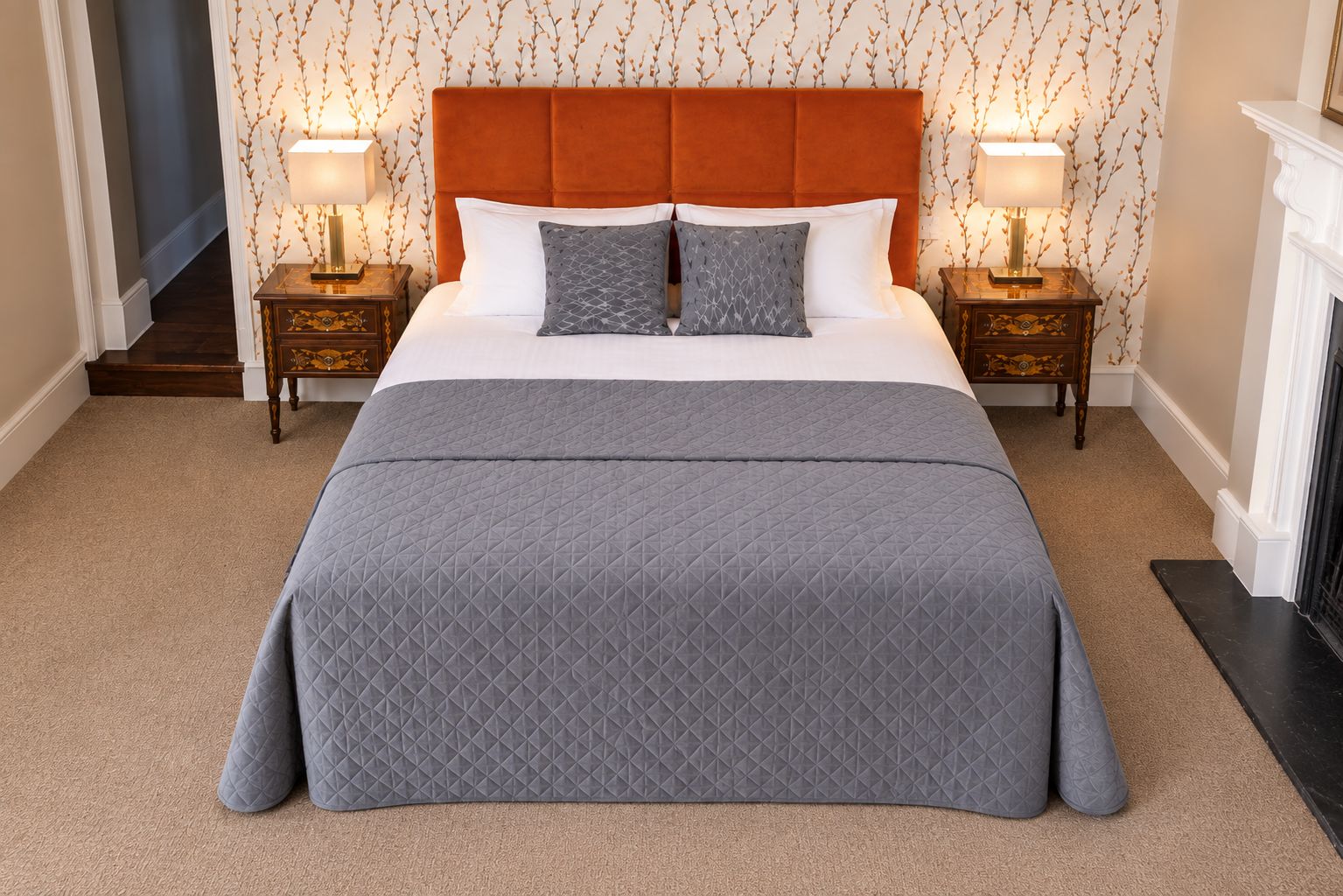 A neatly made bed with a grey quilt, two bedside tables with lamps, and a patterned wallpaper.