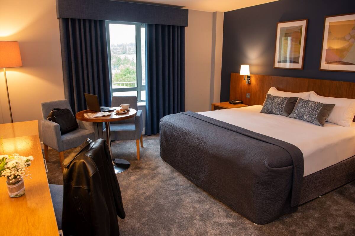 Quarto de hotel com uma cama grande, uma escrivaninha com um laptop, uma cadeira, uma pequena mesa com uma xícara de café e vista para o exterior pela janela.