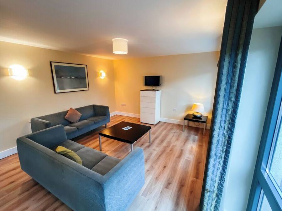 Uma sala de estar moderna com um sofá cinzento, uma mesinha de centro, uma televisão de tela plana e um abajur sobre uma mesinha lateral.