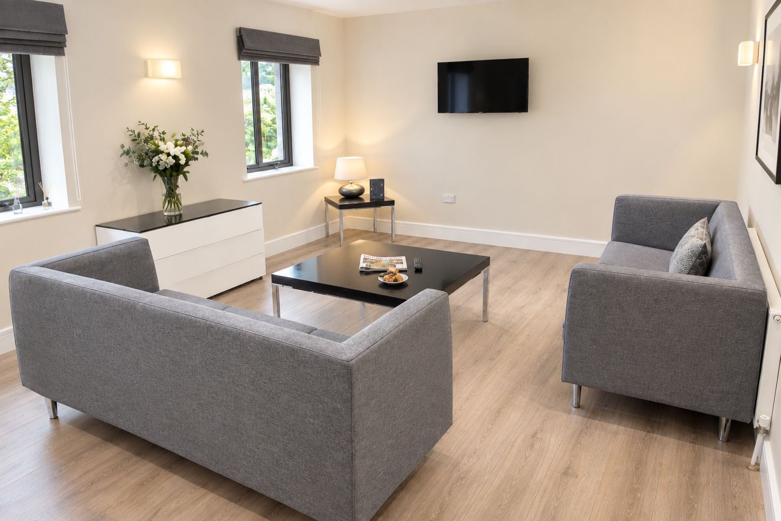 A modern living room with grey sofas, a black coffee table, a TV mounted on the wall, and a window with a view of greenery.