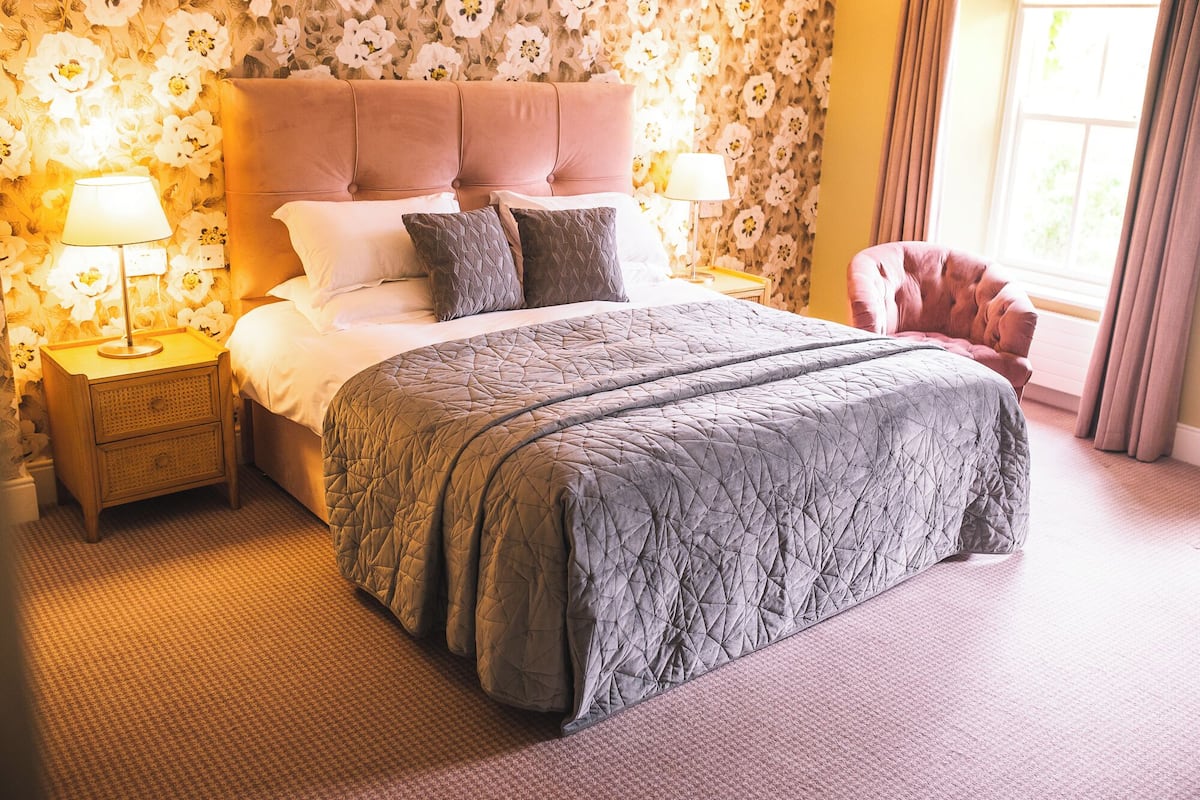 Cama bem arrumada com colcha acolchoada, duas mesas de cabeceira com abajures e papel de parede floral ao fundo.