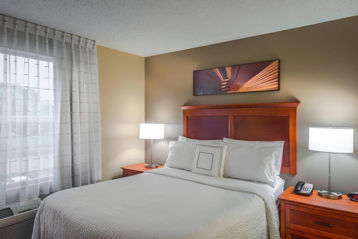 A hotel room with a large bed, two bedside tables, a nightstand, a wall-mounted telephone, and a window with sheer curtains.