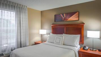 A hotel room with a large bed, two bedside tables, a nightstand, a wall-mounted telephone, and a window with sheer curtains.