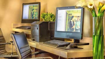 A hotel reception desk with two computer monitors, a keyboard, a mouse, and a chair. There is a vase with flowers on the desk.