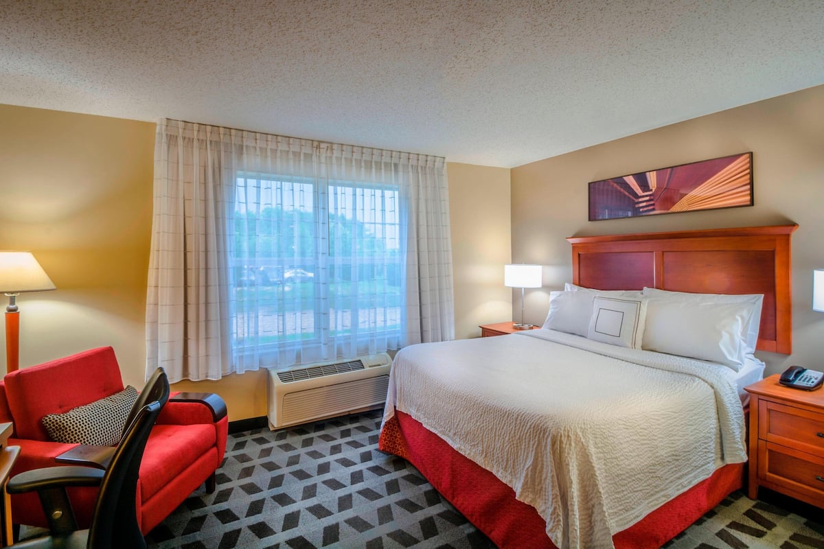A hotel room with a bed, a desk with a chair, a window with curtains, a wall-mounted painting, and a nightstand with a phone.