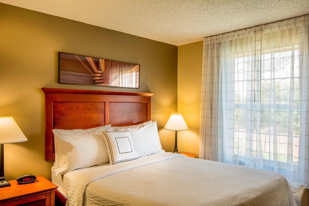 A hotel room with a wooden headboard, a bed with white bedding, two bedside lamps, a digital clock, and a window with sheer curtains.
