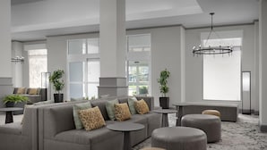 A modern hotel lobby with a sectional sofa, ottomans, round tables, potted plants, and a large window. - Hilton Garden Inn Annapolis (Annapolis)