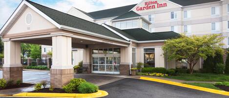 A hotel entrance with a covered waiting area, a clear sky, and a sign for the Hilton Garden Inn.