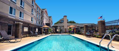 A swimming pool with a clear blue water, surrounded by lounge chairs and umbrellas, and a hotel building in the background.