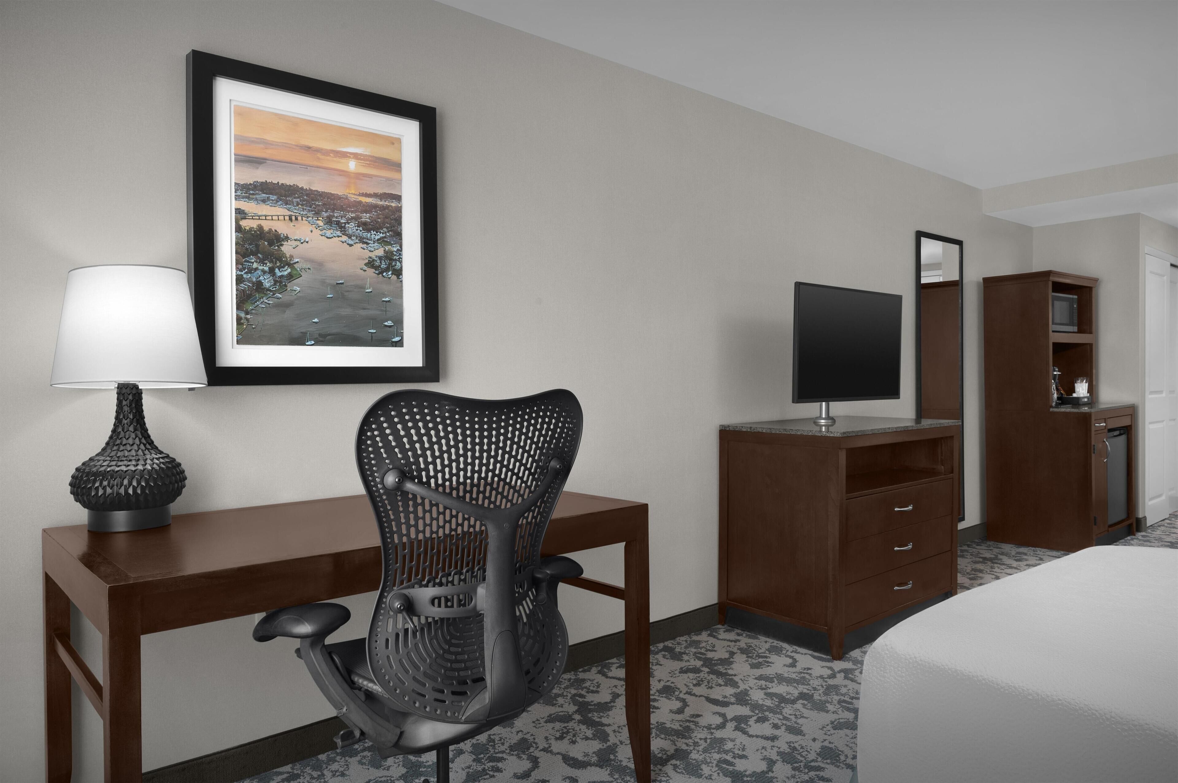 A hotel room with a bed, a desk with a chair, a TV, a mirror, and a framed picture on the wall.