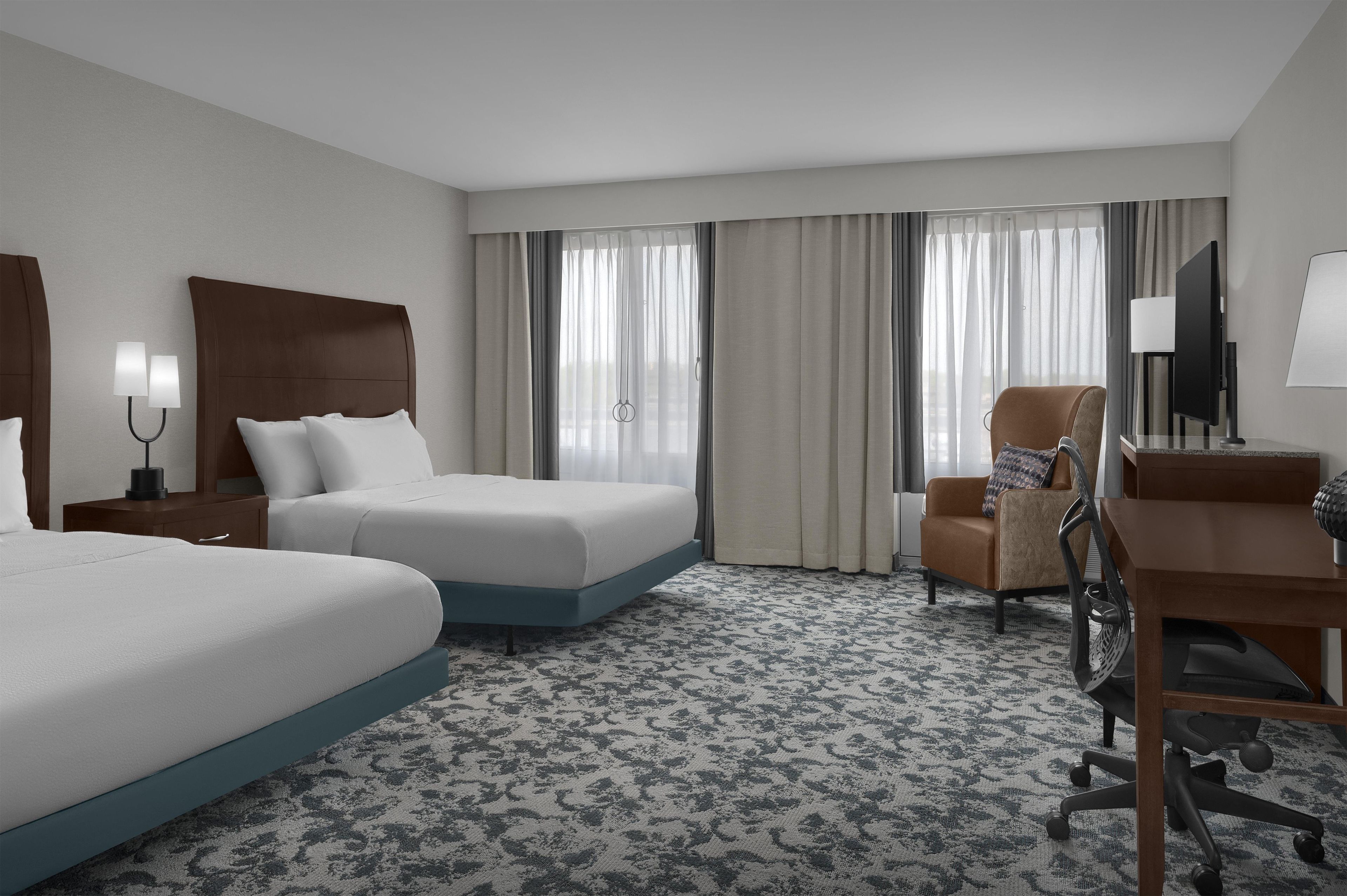 A hotel room with two beds, a desk, a chair, a TV, and a window with curtains.
