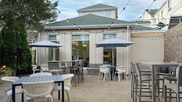 An outdoor patio with tables and chairs, a brick floor, and a building with a green roof in the background.