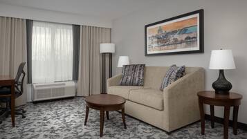 Een hotelkamer met een bank, twee bijzettafeltjes, een lamp, een stoel, een bureau en een groot raam met gordijnen.