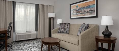 Habitación de hotel con sofá, dos mesitas auxiliares, lámpara, silla, escritorio y un amplio ventanal con cortinas.