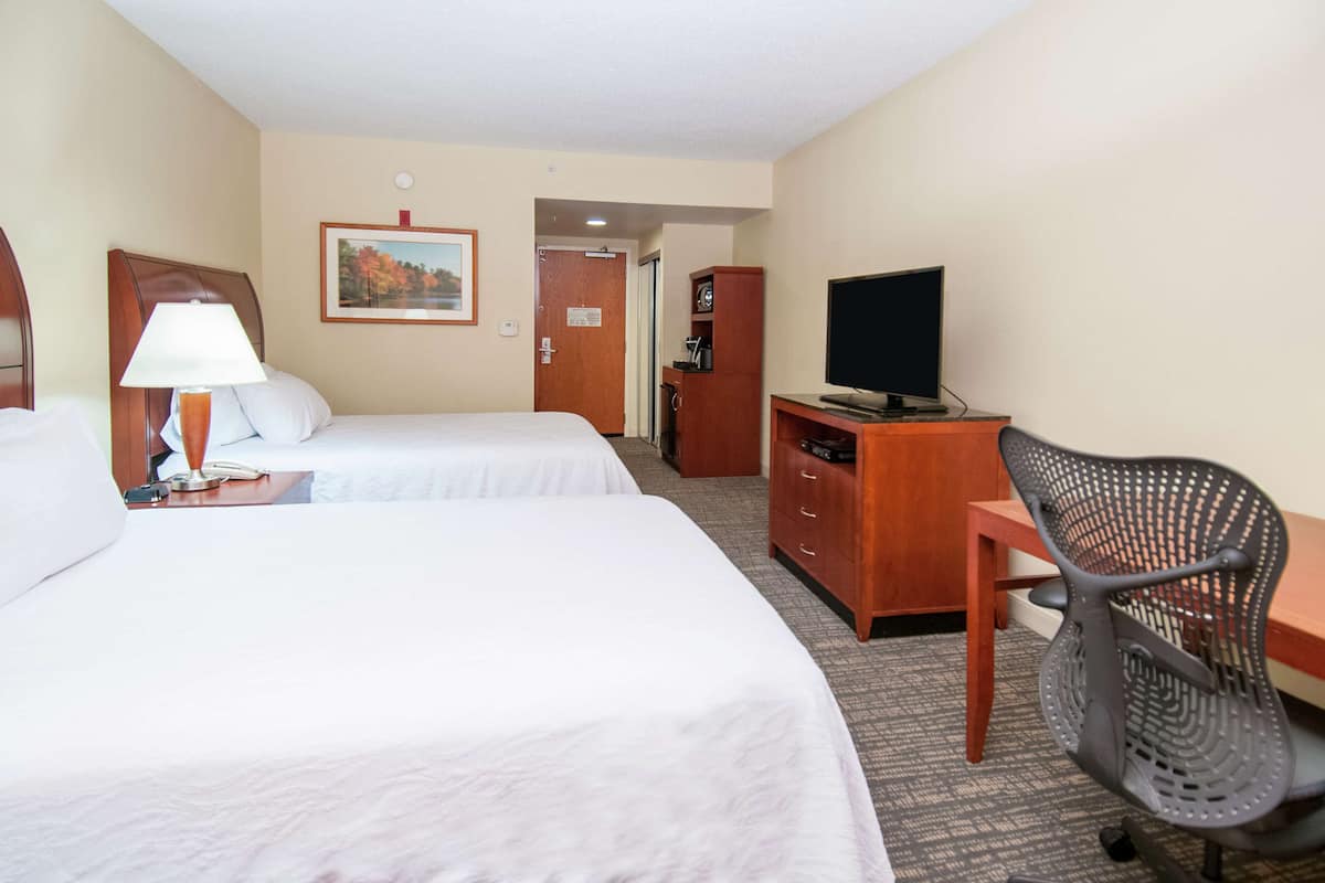 A hotel room with two beds, a desk with a television, a chair, a lamp, and a door leading to another room.