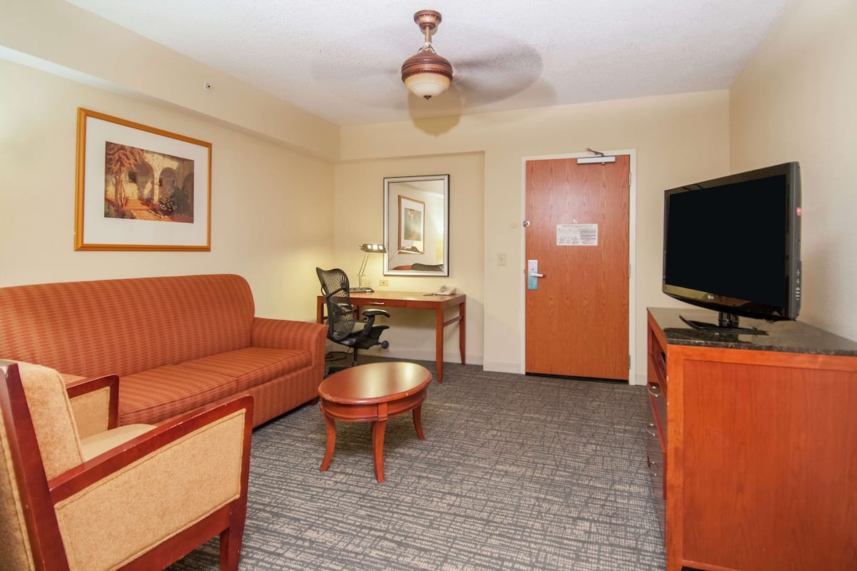 A hotel room with a sofa, armchair, small table, TV, desk, chair, and ceiling fan.