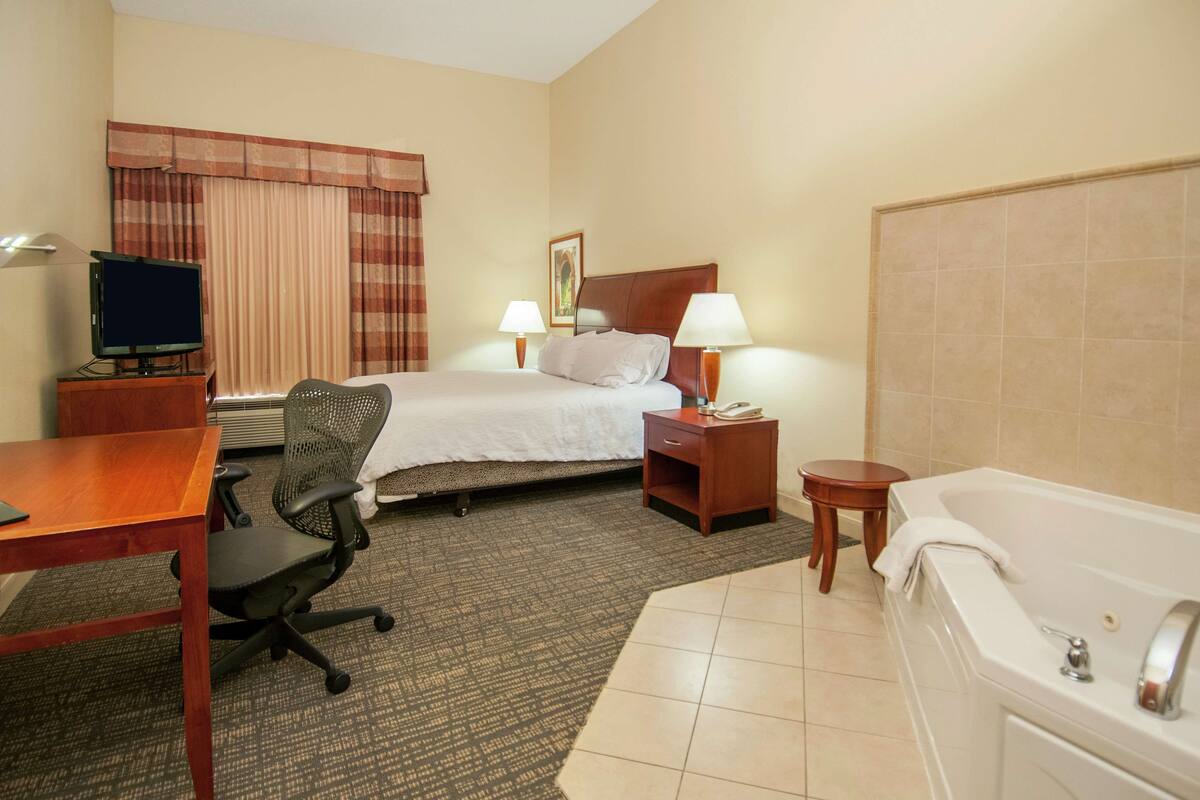 A hotel room with a bed, desk, chair, TV, lamps, and a bathtub.