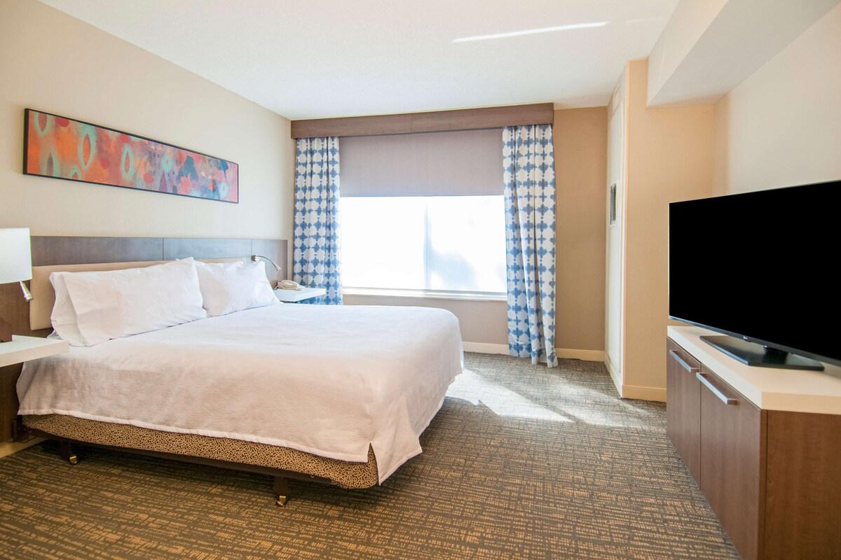 Una habitación de hotel con cama, televisión, escritorio y ventana con cortinas.